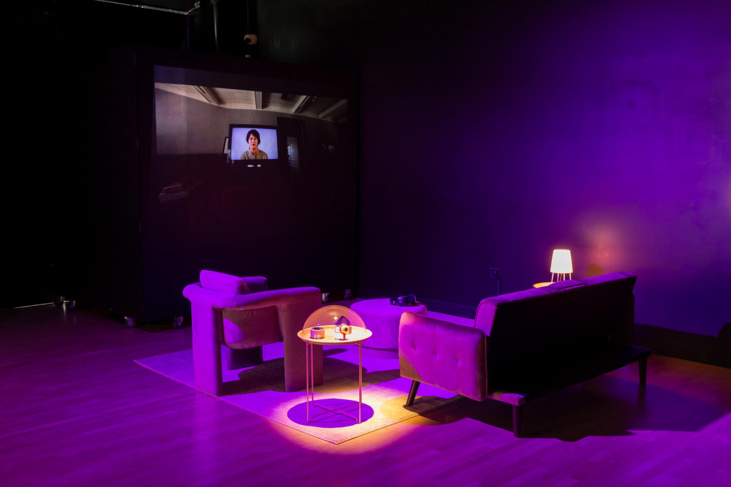 A model of a living room lit purple, with a screen in a high corner showing a woman's face looking down