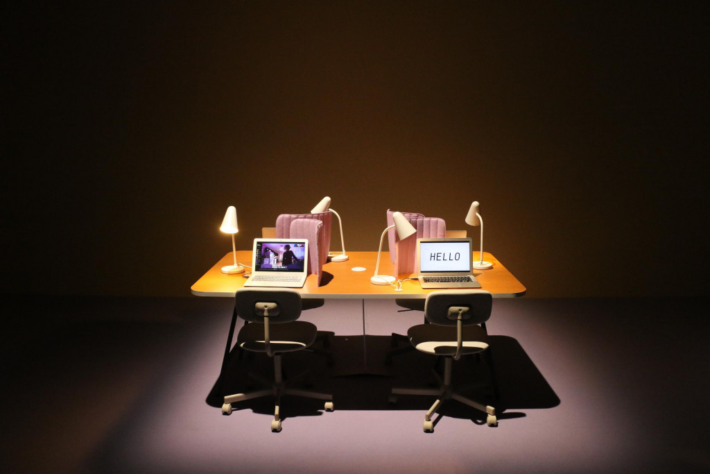 A table spotlit in a dim room. There are laptops on it, with desk lamps and pink cushions that provide some privacy for the user