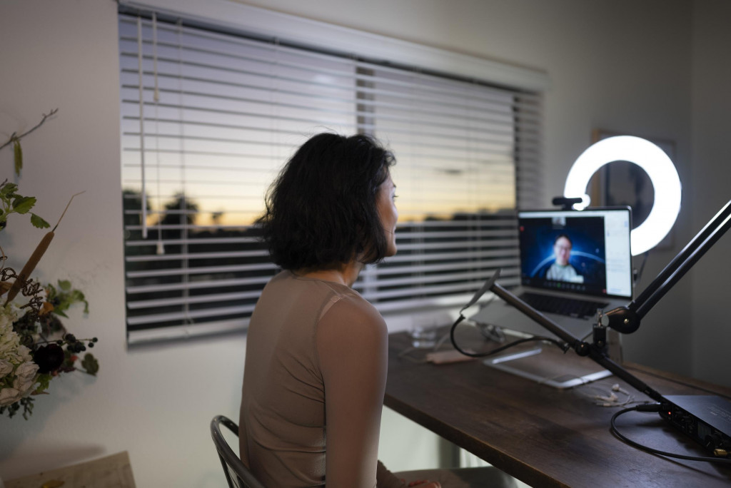 A woman sits at a desk in front of a computer laptop mounted on a metal arm. A ring light illuminates her face and the light in the window is dusky