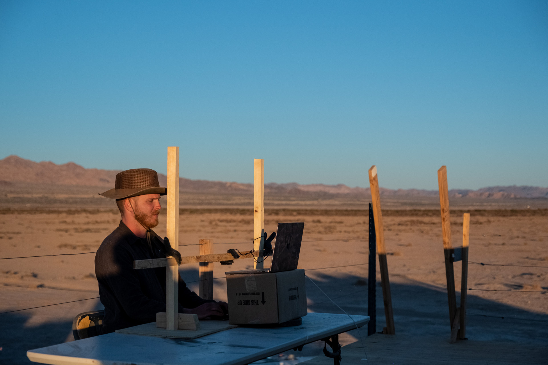 A man in a wide brimmed hat sits at a computer. Behind him is blue sky and desert