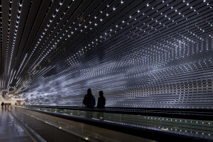 Two people ride a moving walkway in a tunnel illuinated by small white lights