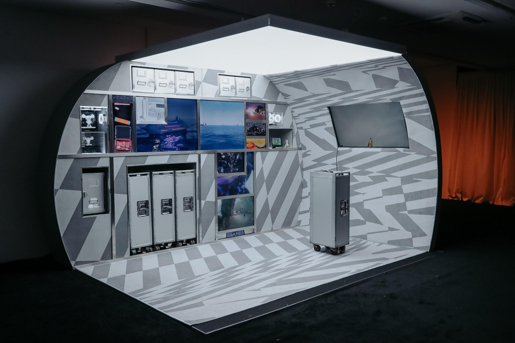 A photograph of a display booth modeled after an airline gallery, with screens nestled among the rolling carts and cabinets showing a variety of digital animations