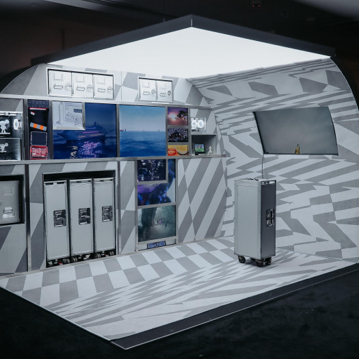 A photograph of a display booth modeled after an airline gallery, with screens nestled among the rolling carts and cabinets showing a variety of digital animations