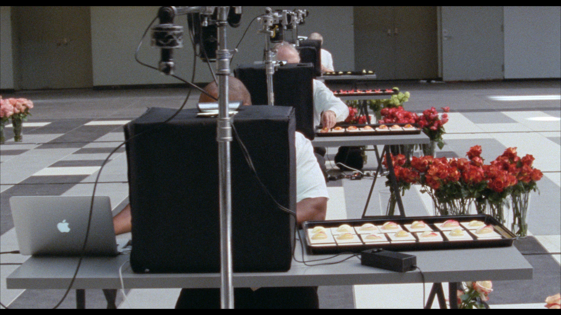 A film still showing men seated at desks with computer equipment, vases of roses, and petals laid out on sheets of paper in trays