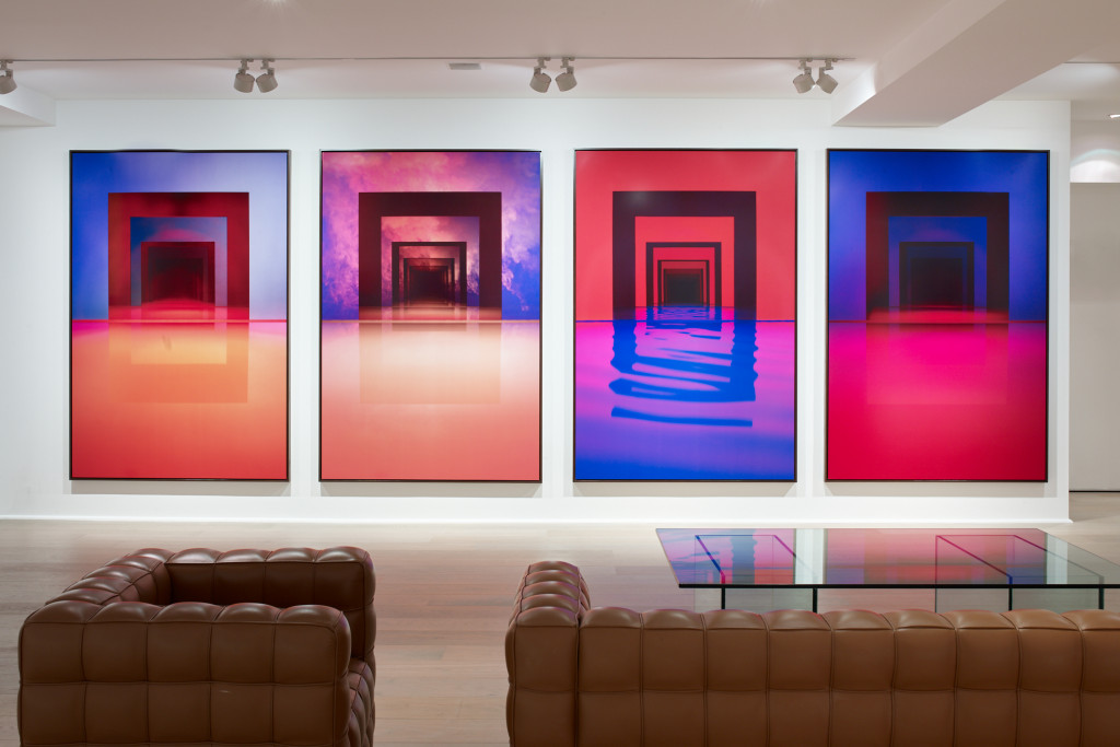 Four large-format photographs hang in a gallery near large brown leather sofas. The images, in rich tones of pink, purple, and blue, show square gates over reflective pools