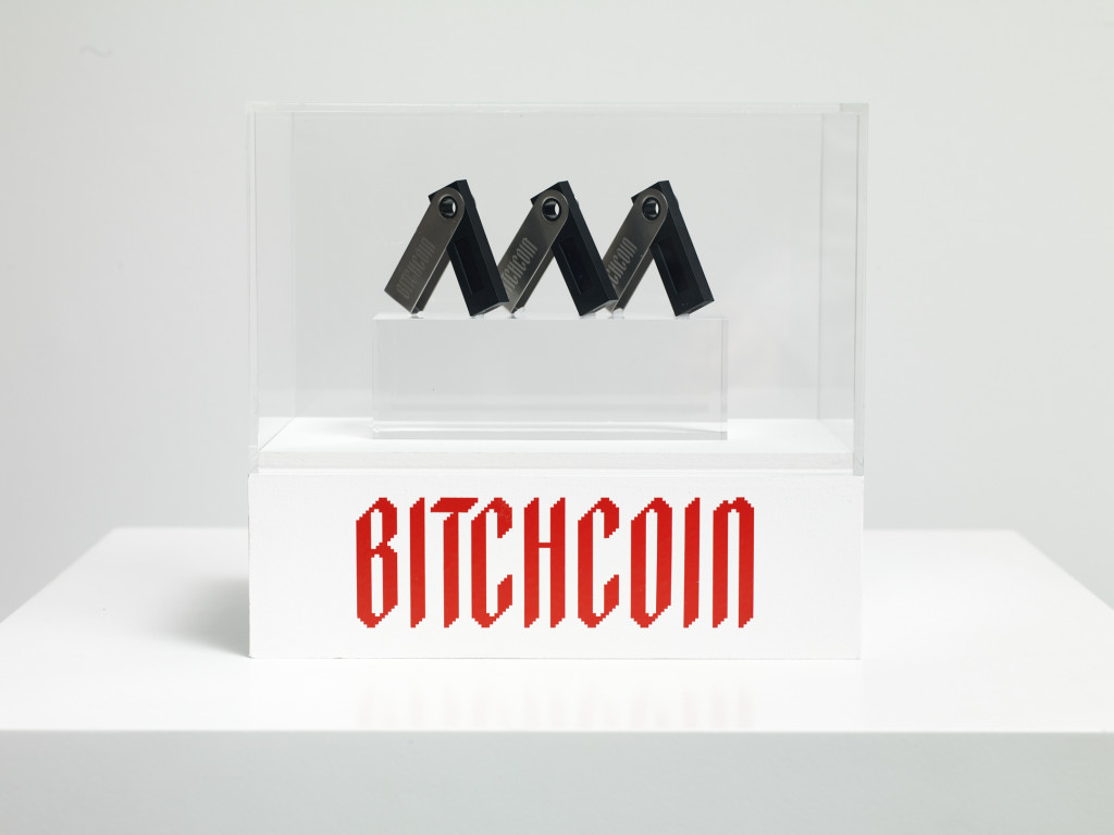 A plexi box on a white plinth holds three black thumb drives. Red text on the plinth reads Bitchcoin