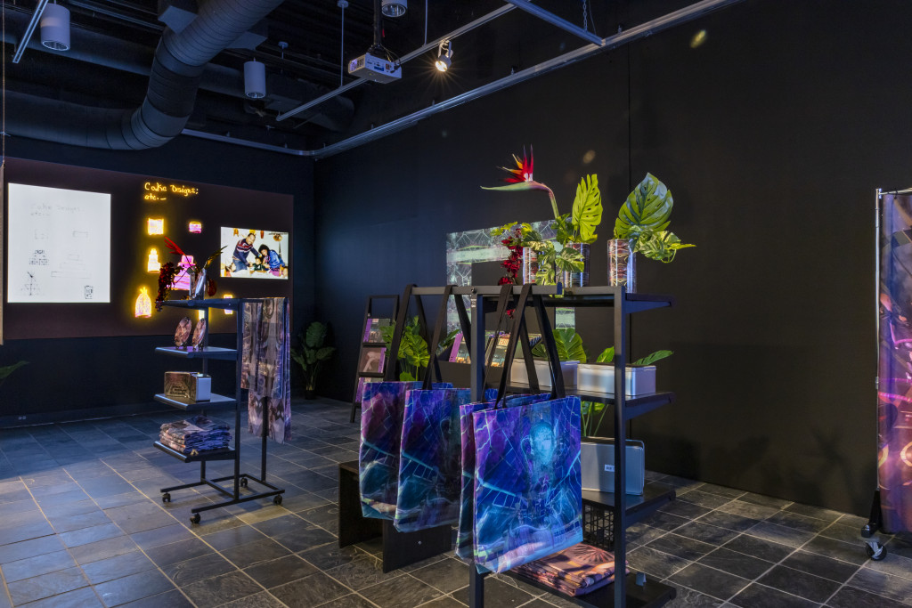 A black-walled gallery with shelves displaying plants and printed textiles