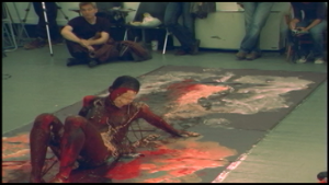 A grainy video still of an artist dragging his body across a canvas on the floor; both he and the fabric are covered with red and white pigment