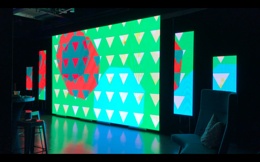 A digital abstract image comprising white triangles and overlapping blue and red octagons on a green field is projected across several large screens in a dark room