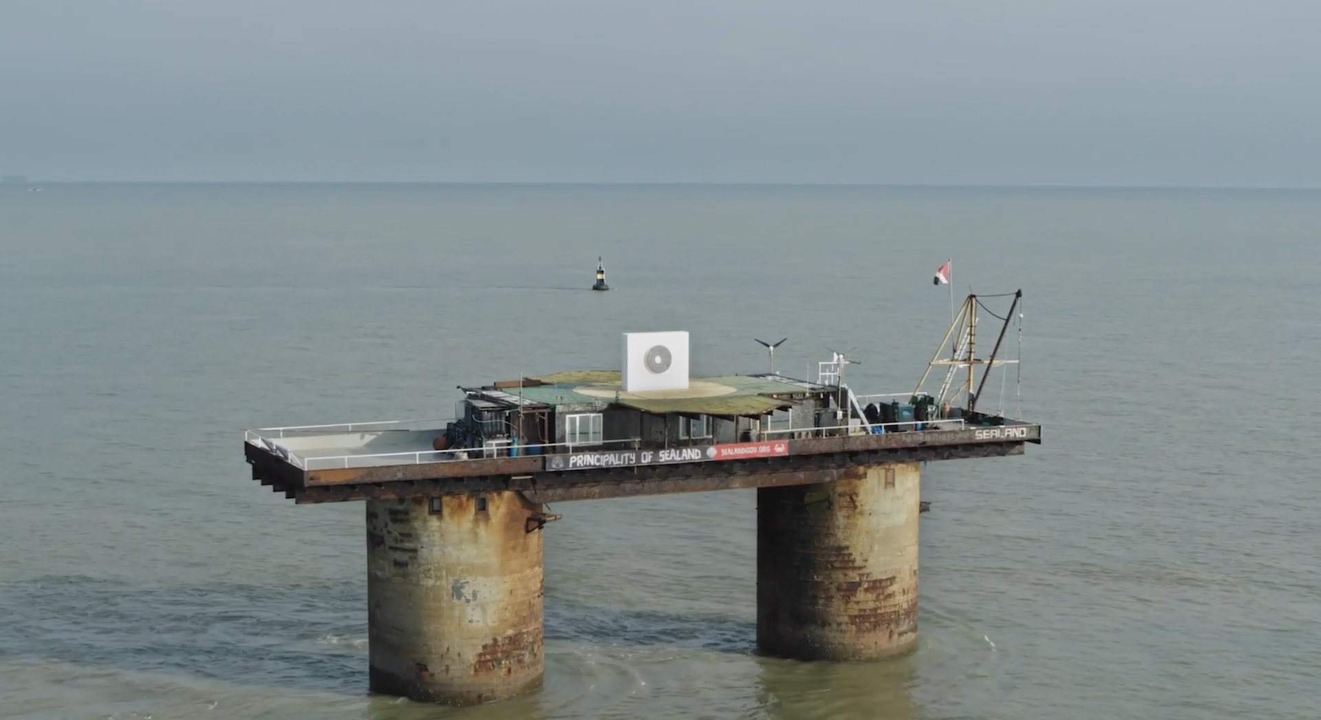 A distant view of a round, gray painting hanging on a white temporary wall, positioned atop a helipad on a platform in the ocean