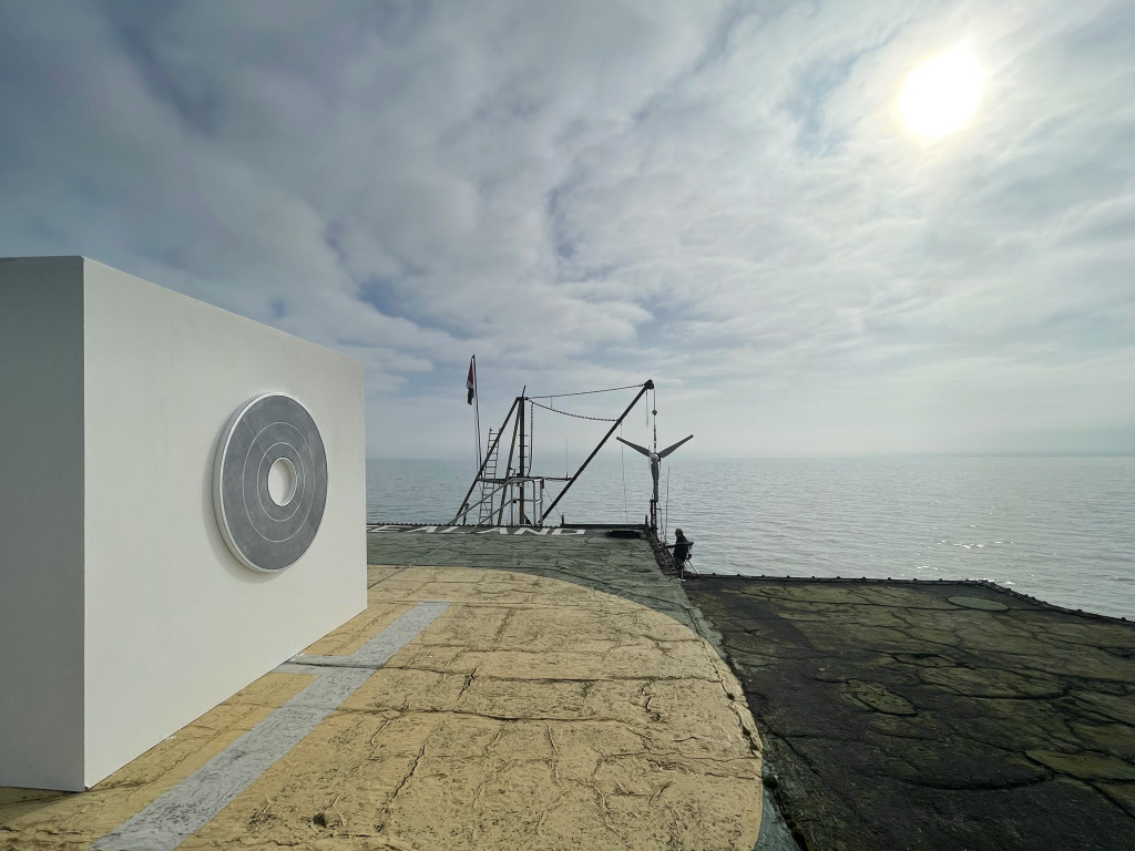 A gray round painting hangs on a white wall atop a weathered concrete platform in the ocean