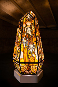 A crystal-like column with facets stained-glass depicts a female figure, looking downward and holding drooping flowers