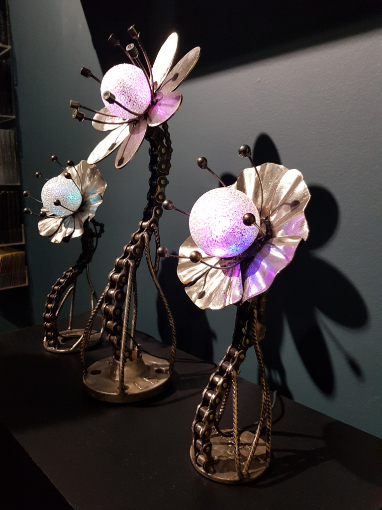 Three small, sinewy metallic sculptures of flowers line up against a gray wall