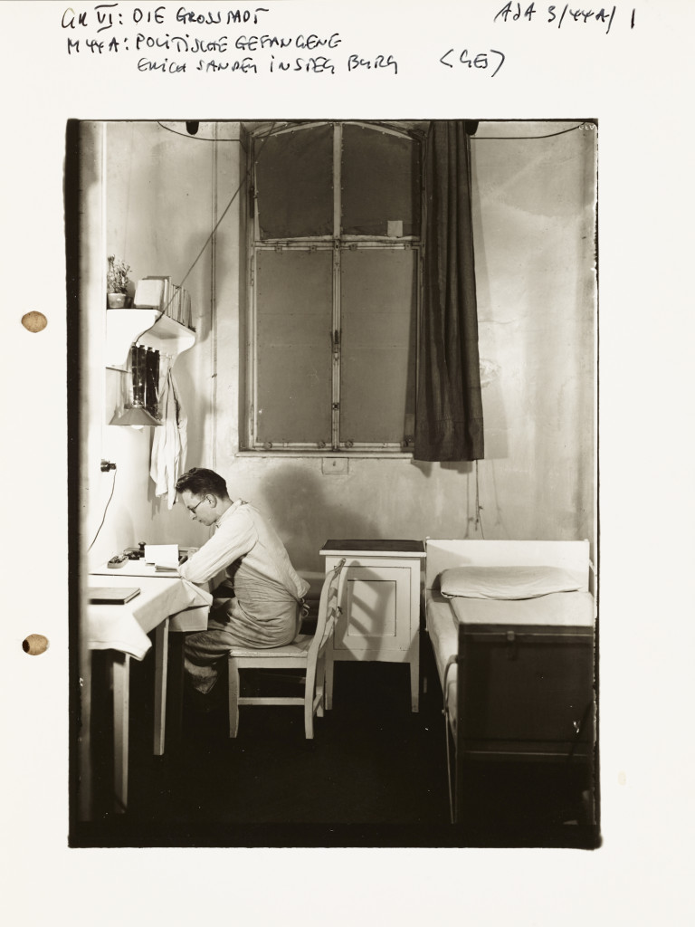A black and white photo of a man reading at a desk in a narrow, sparely furnished room. There are wide margins around the photo, with handwritten notes at the top