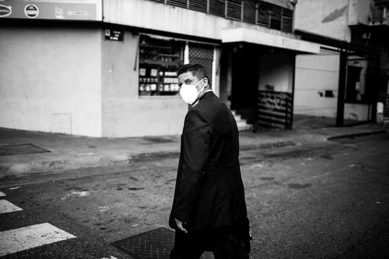 A black-and-white photograph of a man in a dark suit and a white face mask, turning over his shoulder to look at the photographer as he crosses the street