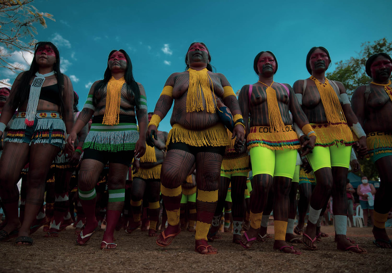 Five dark-skinned women wearing bright beaded fringe around their necks and waists stand in a row holding hands