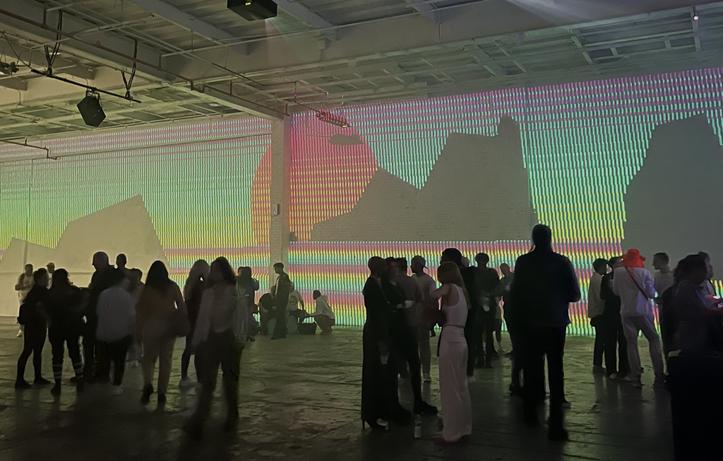 People in groups stand in a large room. Behind them is a large projection of a low-fi, technicolor landscape