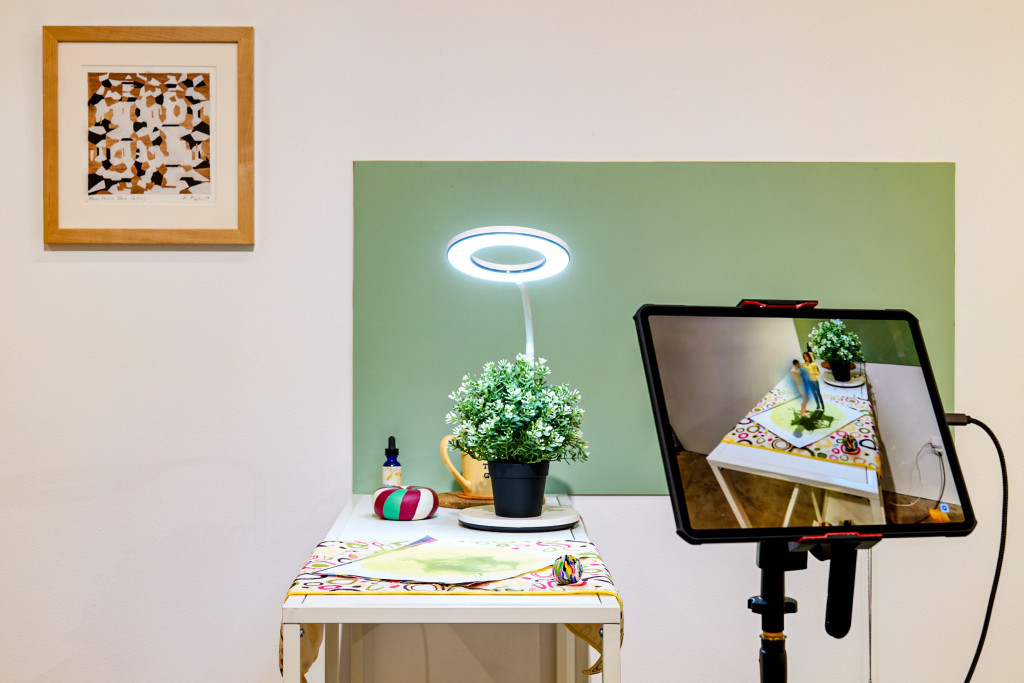 A photo of an installation, where a potted plant sits with other objects on a shelf under a ring light. A tablet next to the assemblage shows it with the addition of animated figures