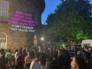 At dusk, people gather outside a stone building that has song lyrics projected onto its exterior