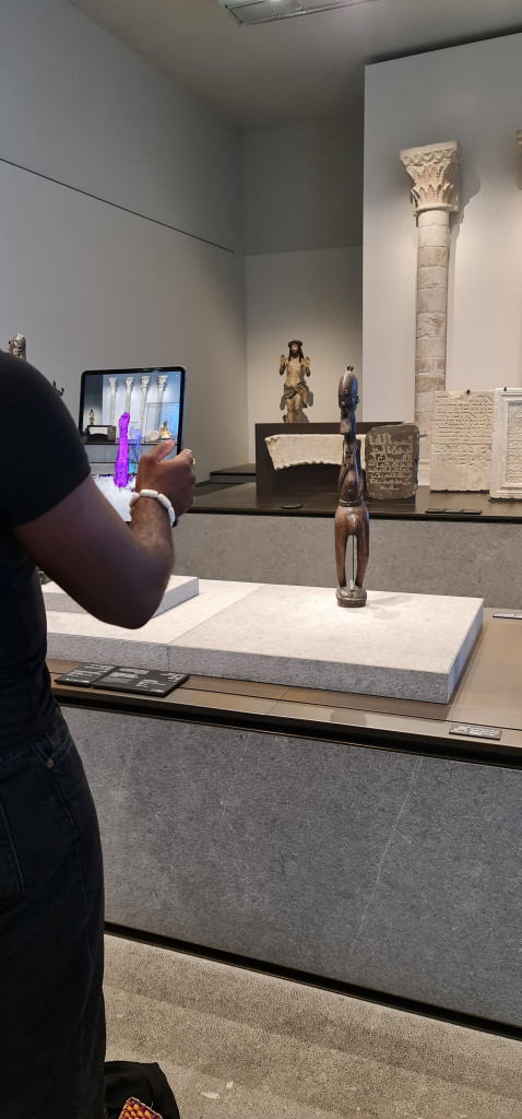 A man holds up a tablet screen in front of a Benin bronze sculpture in a museum
