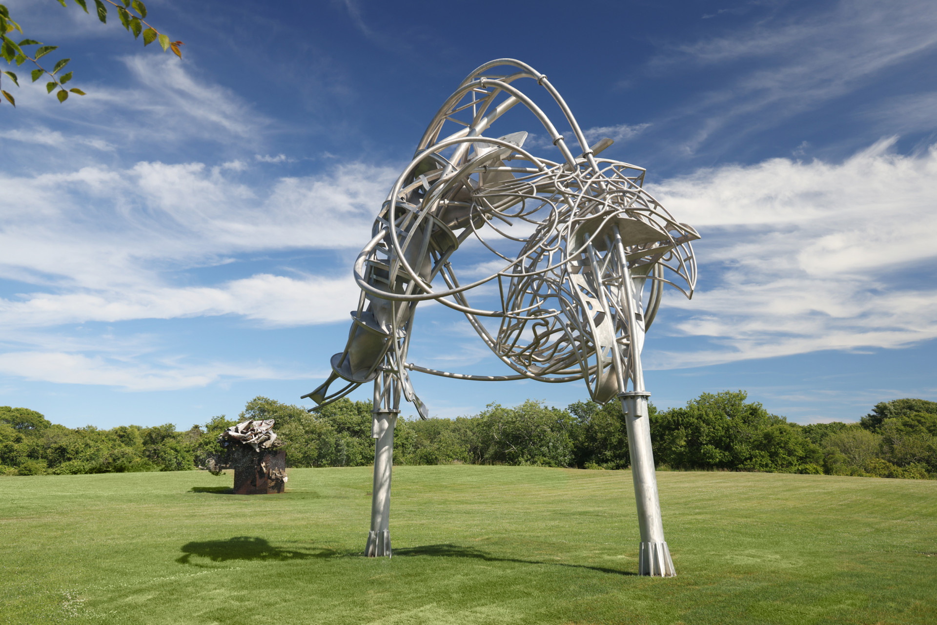 A photograph of an arcing metallic sculpture with swooping, curving rods, standing in a green field under a blue sky