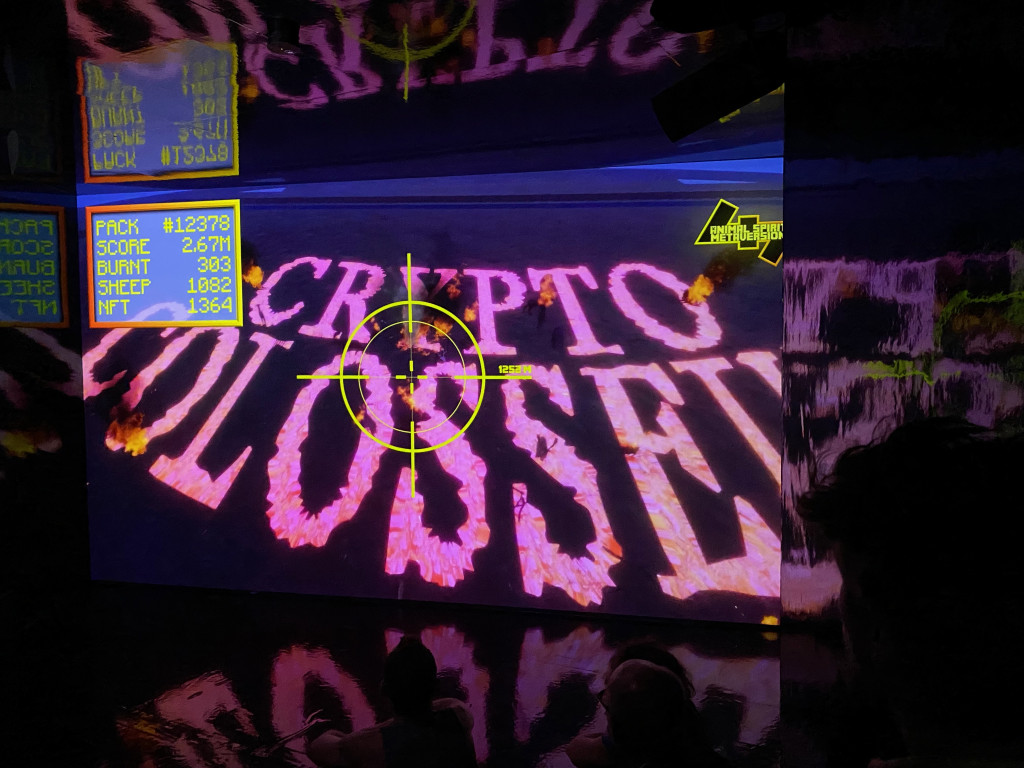 A video projection on a wall shows a yellow target over the words "Crypto Colosseum" in pink lettering, with a video game score chart in the upper left corner