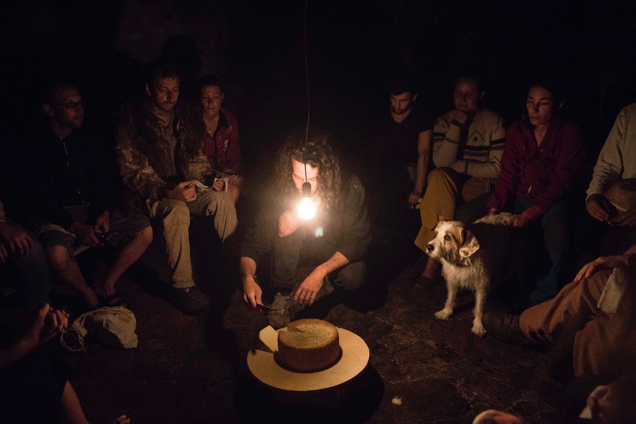 A group of people gathers in a dark room around a man cutting into a circle of cheese