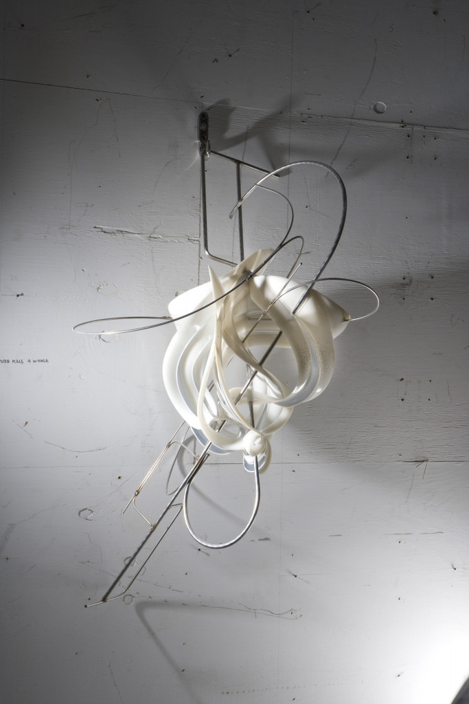 A 3Dprinted sculpture of diaphanous white ribs affixed to curving metallic rods hanging on a wall in a studio
