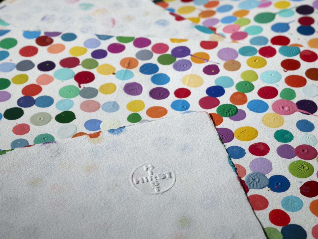 A sheet of paper painted with multi-colored polka dots, overlaid with another sheet of paper showing an embossed stamp spelling "HIRST"