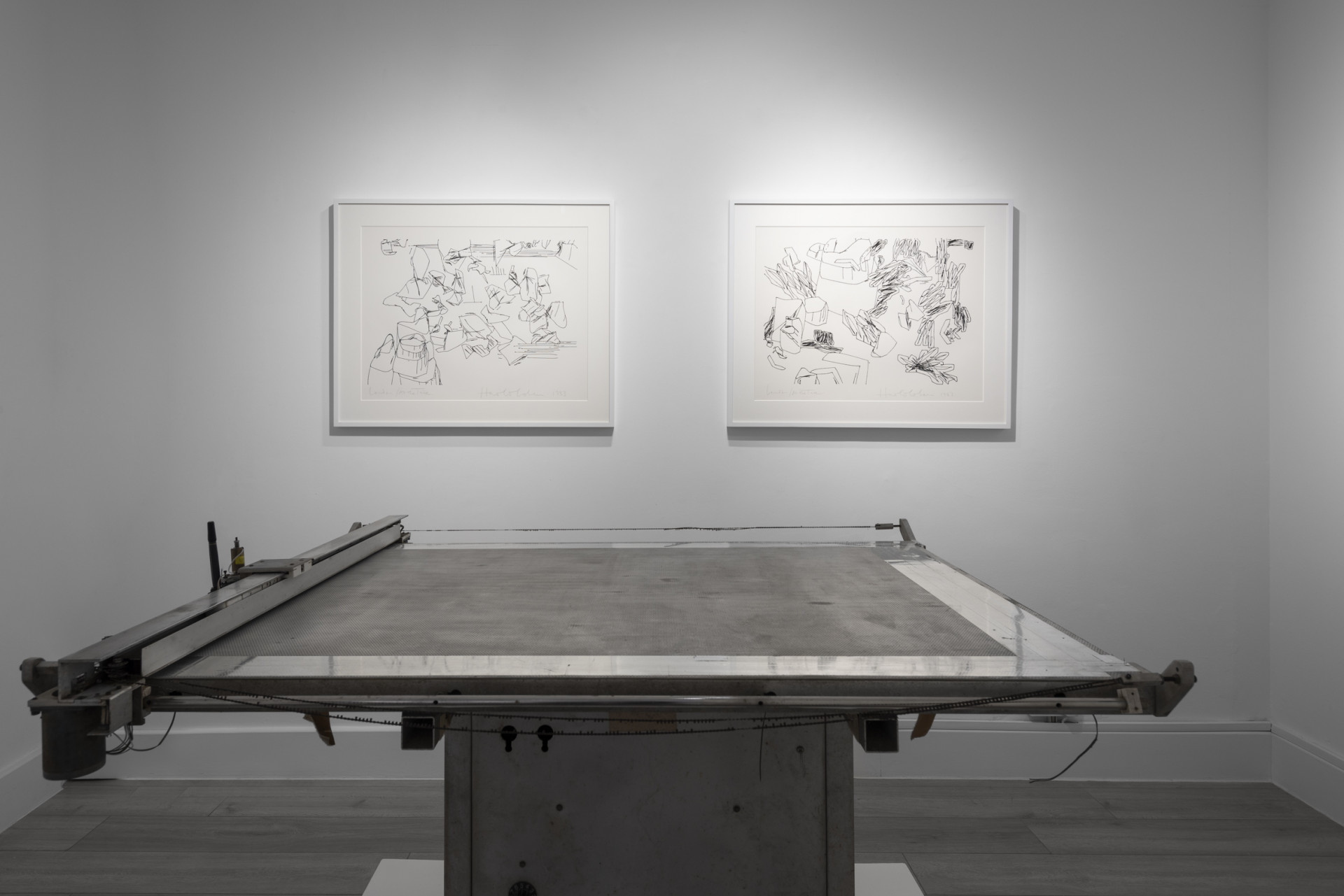Install shot of a white cube gallery with a grey drawing machine in the foreground and two framed black-and-white doodles hanging on the wall behind