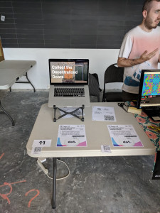 A photograph of a folding table presenting a digital collectible project with a laptop and flyers