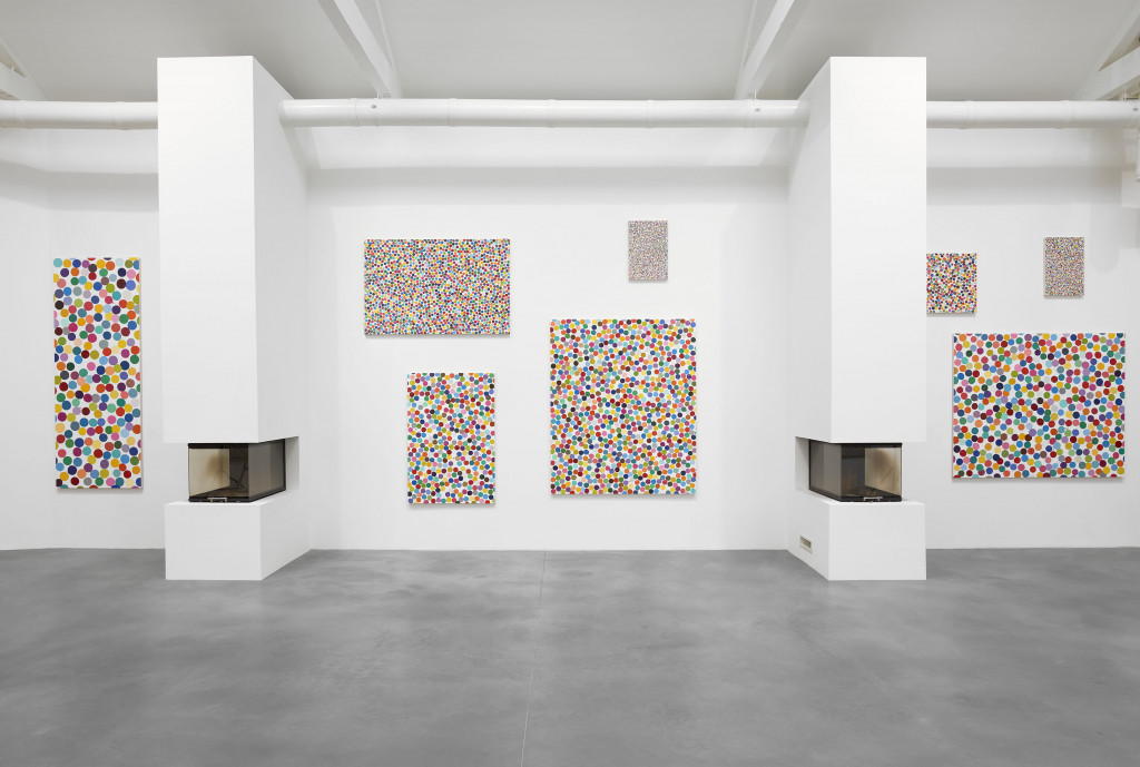 A view of a gallery with multicolored dot paintings on canvas hanging on white walls; there are outcroppings in the walls containing furnaces for burning works on paper