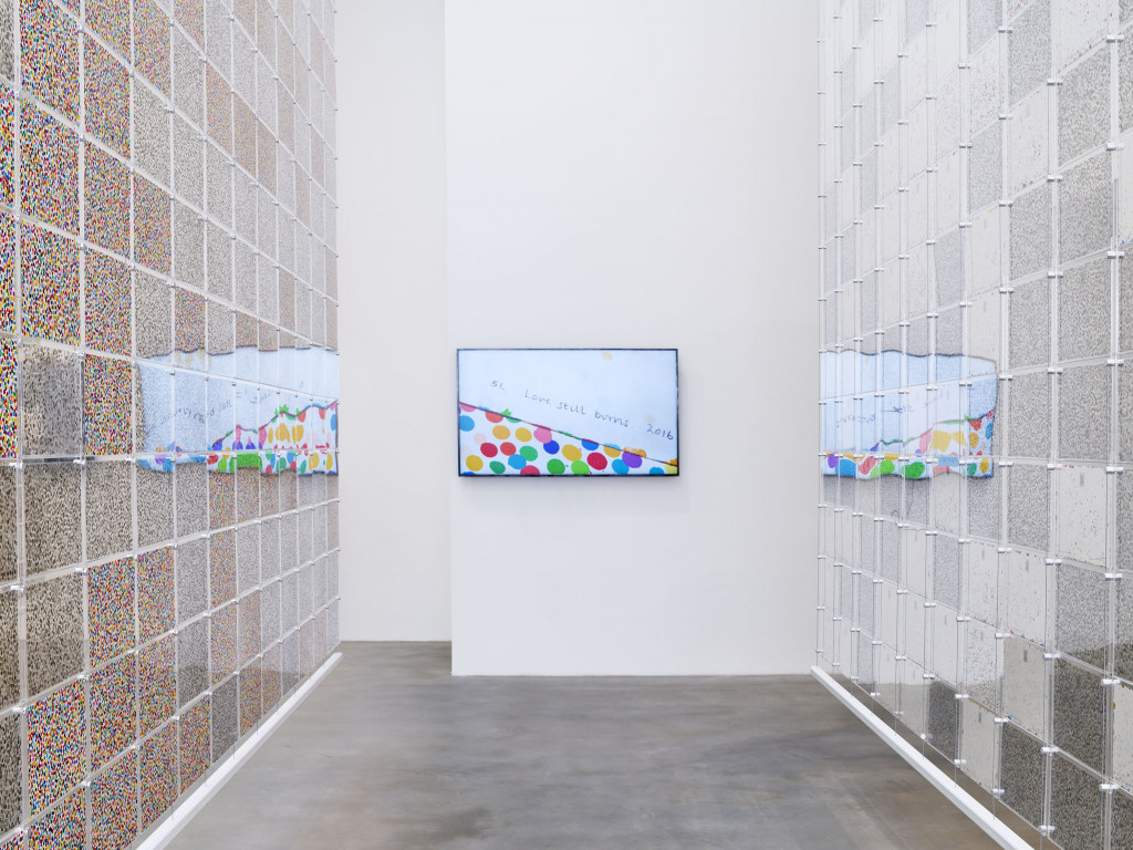 A view of a gallery with a monitor hanging on a white wall; the image on its screen of a multicolored dot painting reflects on two transparent hangings containing more dot paintings