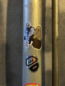 A photograph of a sticker affixed to a metal pole; the sticker depicts a bearded man in a hoodie squatting beside a stone statue