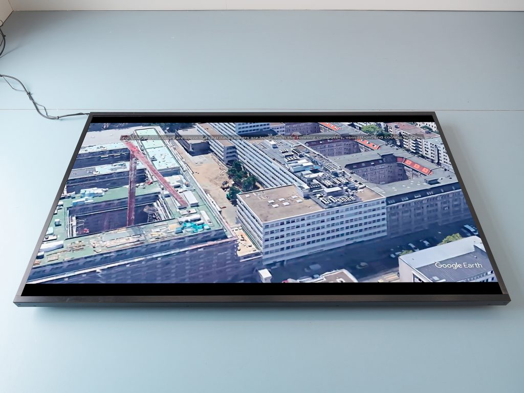 A widescreen TV on a floor showing an aerial shot of a data center on Google Earth