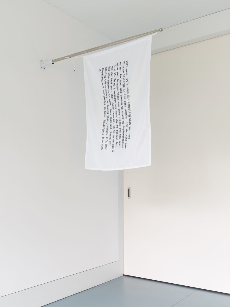 A white cube gallery with a white flag hanging from a rod which is horizontal to the floor; the flag is printed with black text, which is too small to read