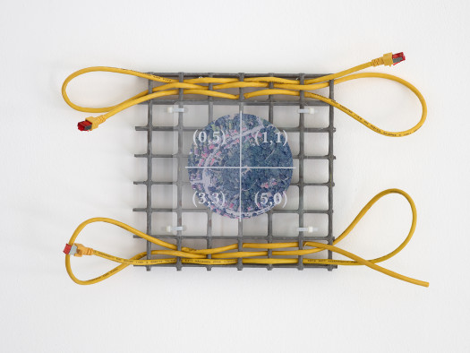 A small grey metal gridded square interwoven with a yellow Ethernet cable, overlaid with a plastic sheet showing an aerial view of a suburban landscape, hung on a white gallery wall