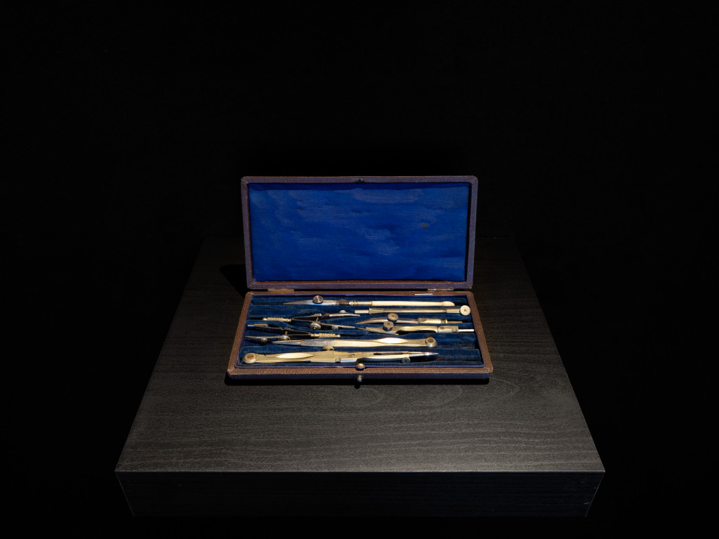 A set of compasses in an open box lined with blue fabric, placed on a black wooden plinth