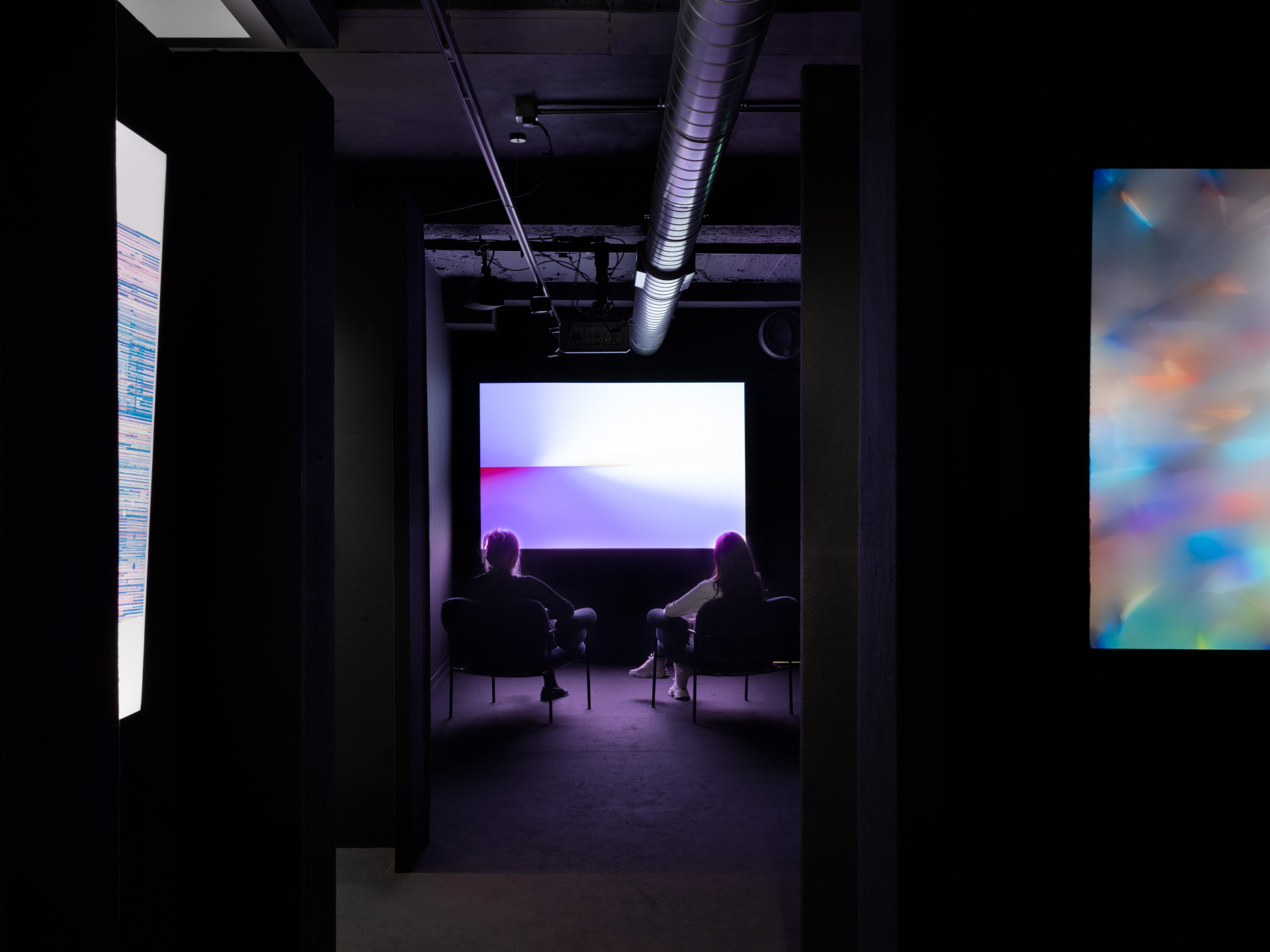 A photograph of a dark gallery with black walls. Two works on screens are partially visible on either side, and a projection of a purplish white abstraction lights up the far wall