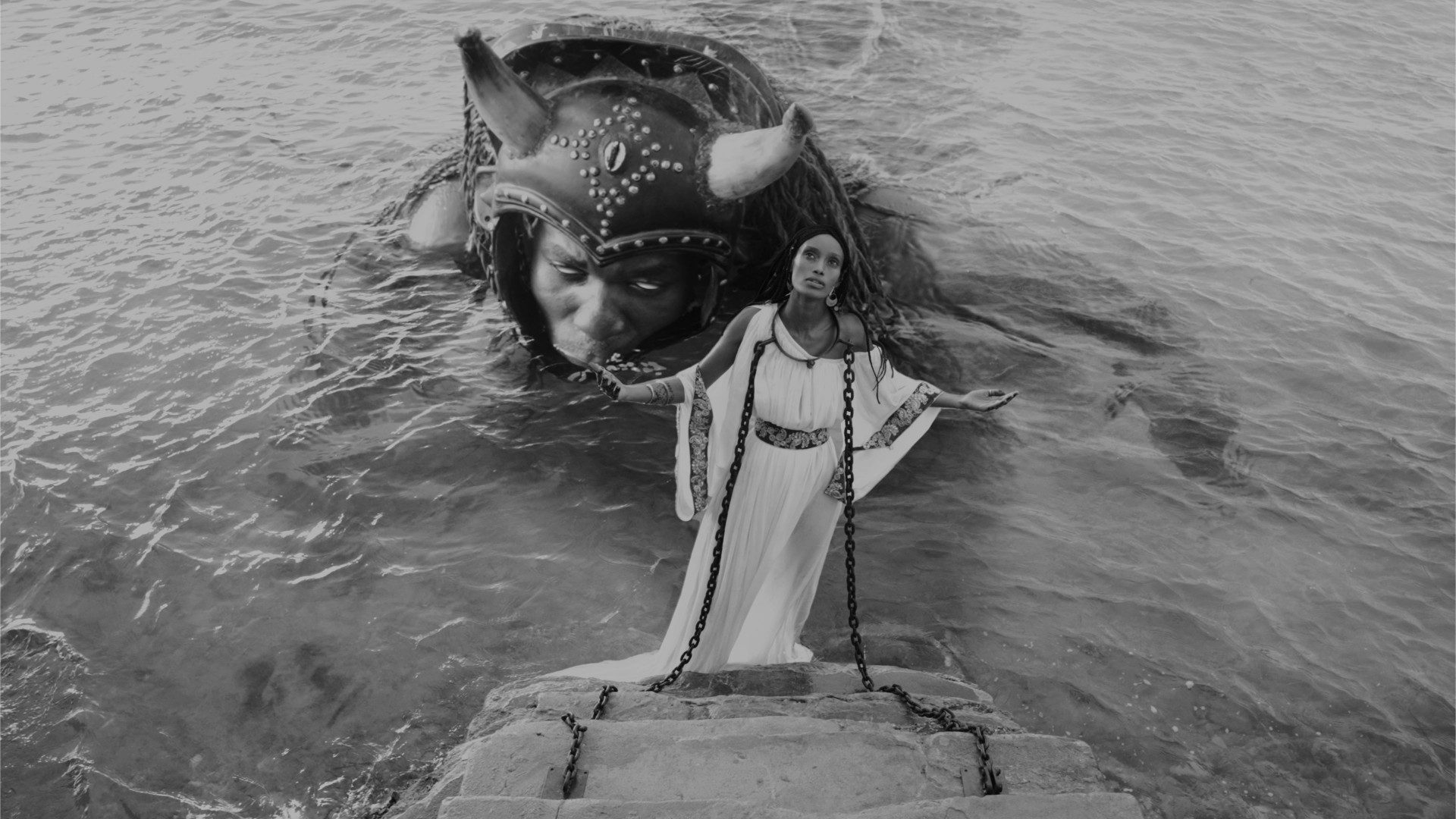 A black-and-white photograph showing a stormy sea, in which a woman stands chained to a rock while a monstrous creature with a human face and horns looms behind her