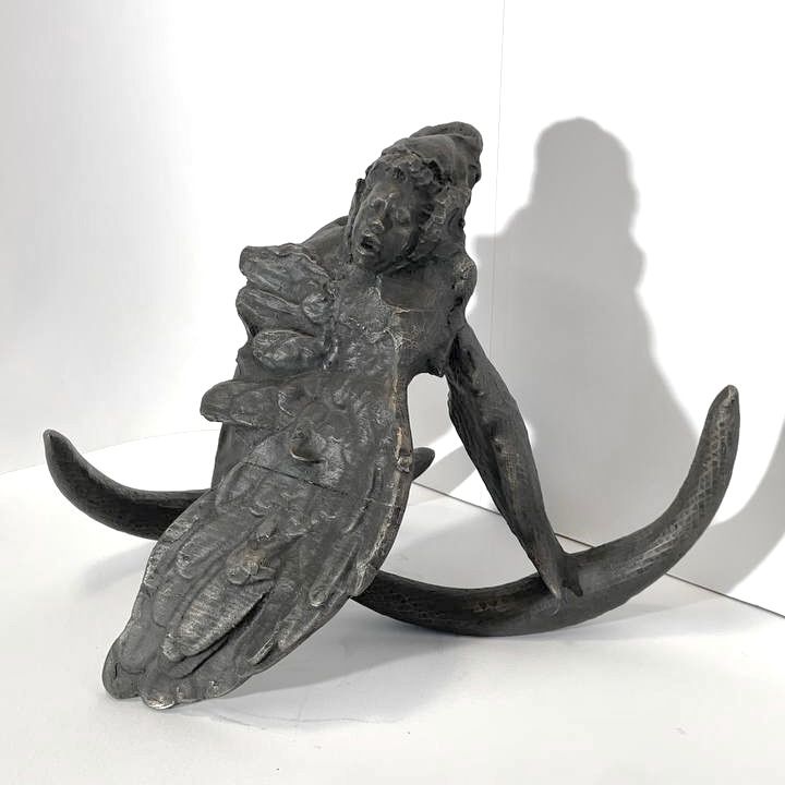 A strange bronze sculpture consisting of a large wing, a woman's head, and two thin crescent shapes, cast in bronze, in a white space