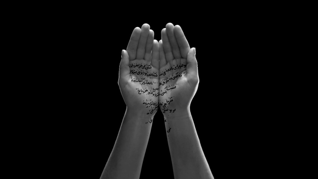 A pair of hands opening to reveal Arabic calligraphy inscribed on the palms, softly lit against a black background