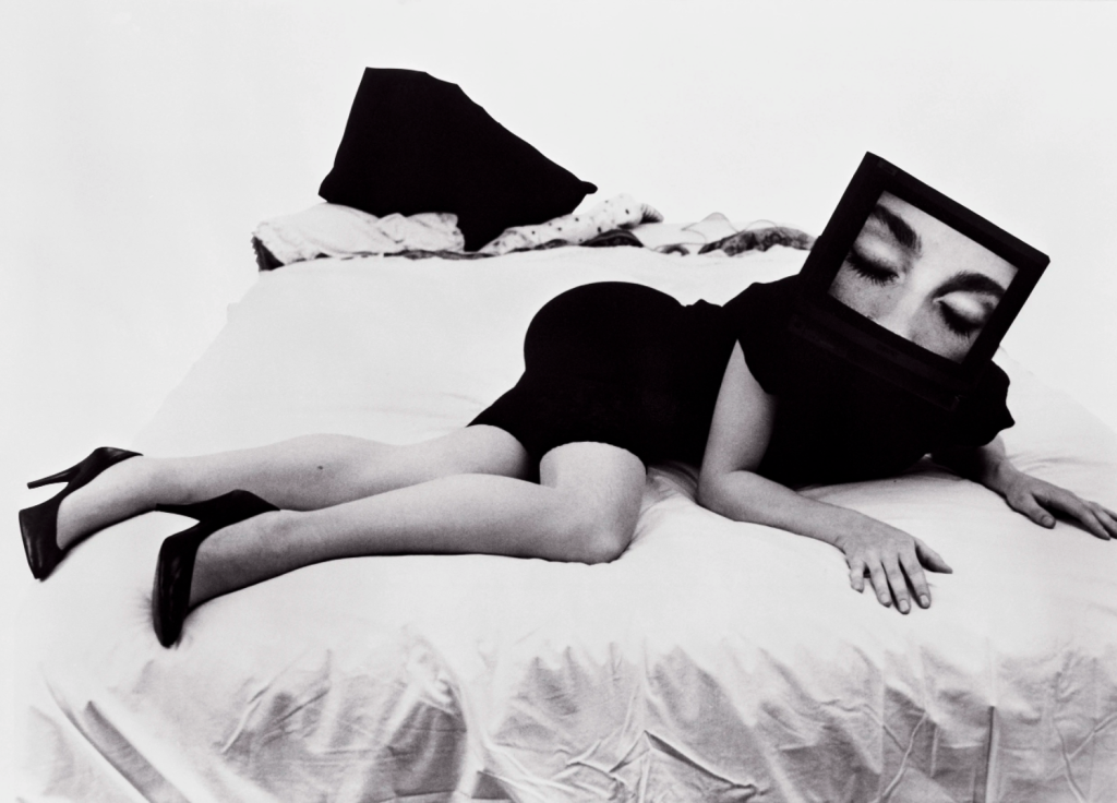 A black-and-white photograph of a woman seductively lying on a bed. Her face is encased in a TV monitor