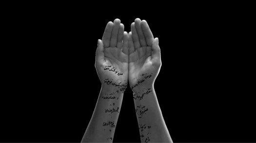 A pair of hands opening to reveal Arabic calligraphy inscribed on the wrists and forearms, softly lit against a black background