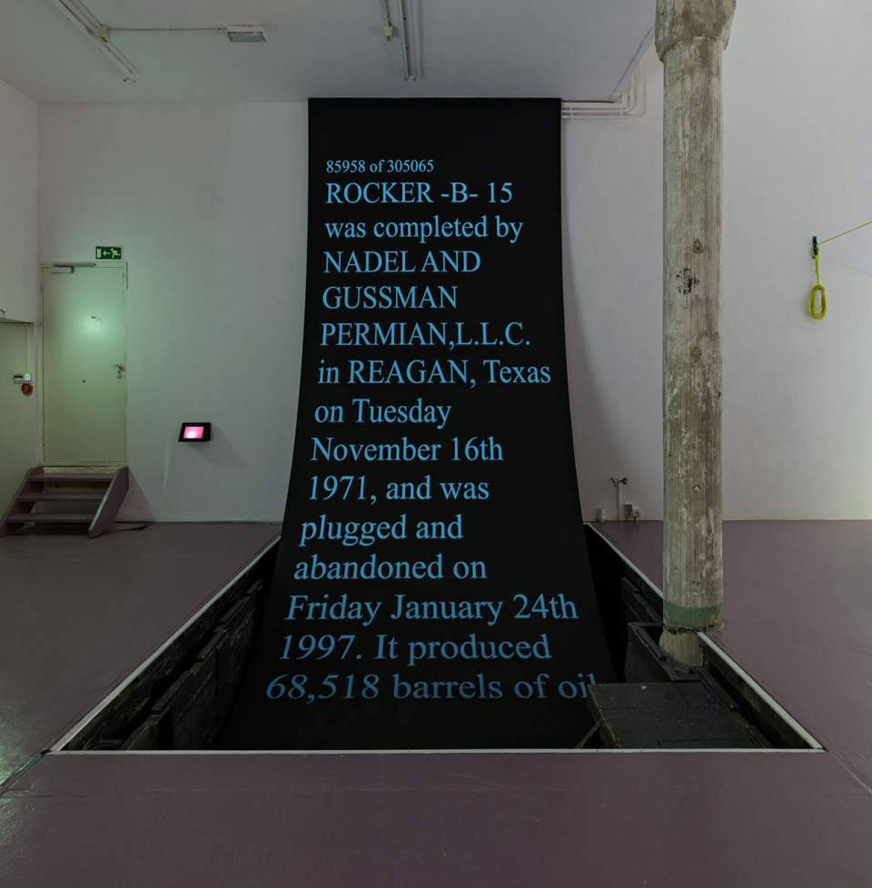 A gallery installation with blue text scrolling up a black sheet that drapes into a hole in the floor