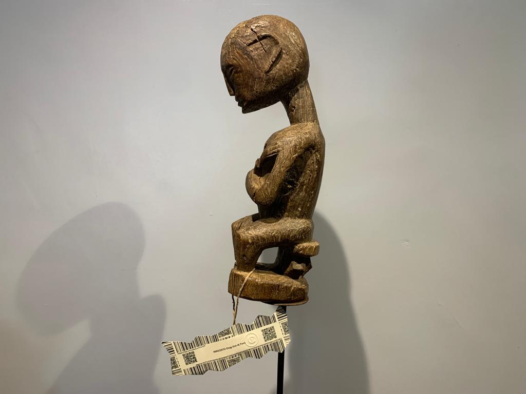 A wooden African statue of a male figure on a stool, with a printed label attached