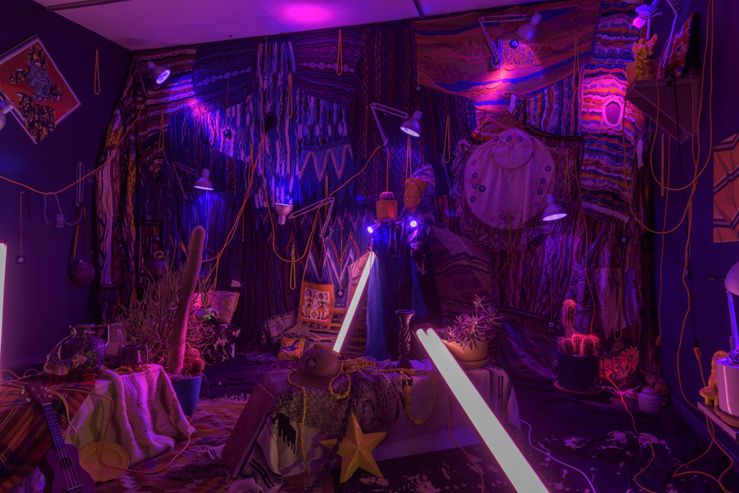 A photograph of a room lit with sparse violet lights and filled with textiles, paintings, and other various clutter