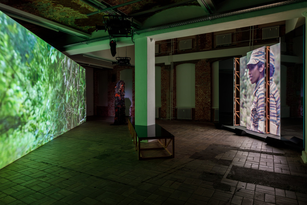An installation view of a gallery space bisected with a white column, with screens on either end of the room, a green jungle scene on the left and a woman in military fatigues on the right