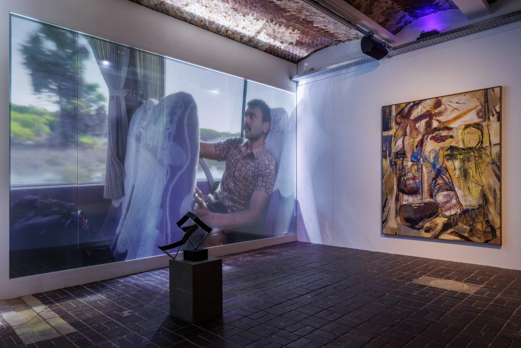 A photograph showing an installation in a gallery, featuring a projected film, a painting, and a sculpture on a plinth