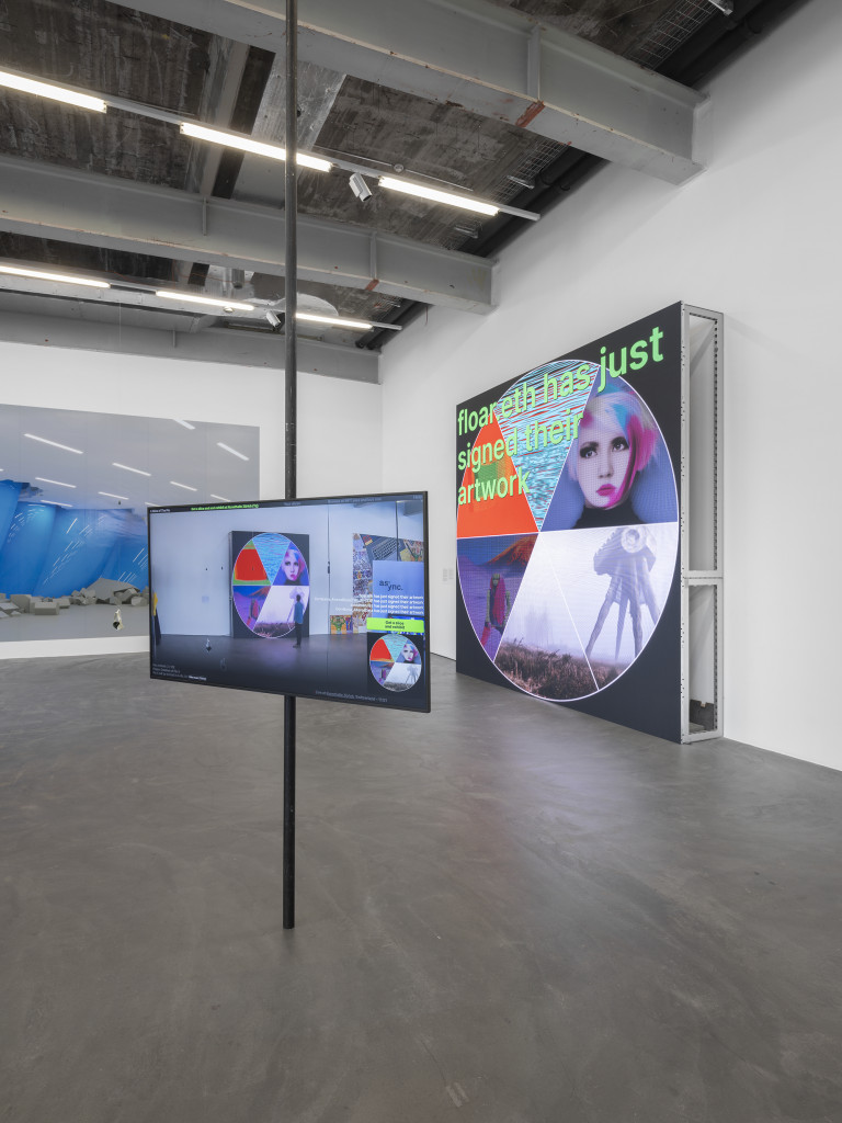 A photograph of an installation in a gallery, showing a projection on a panel with a circle divided into six segments, each showing a different work; in the foreground, a screen mounted on a pole shows the same view with metadata